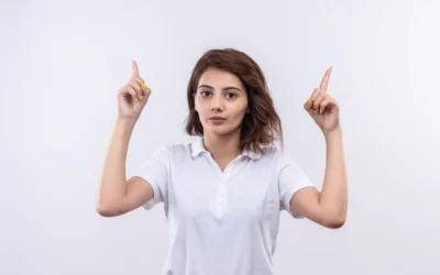 Person in white shirt pointing upwards with both hands against a plain background.